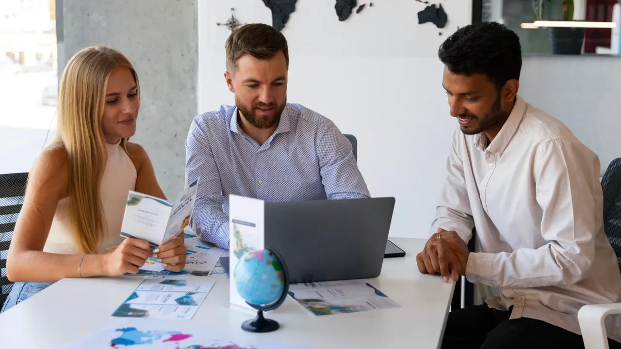 Three colleagues around an office desk reviewing printed comparison sheets and a laptop while shortlisting the best SaaS SEO agencies for 2026