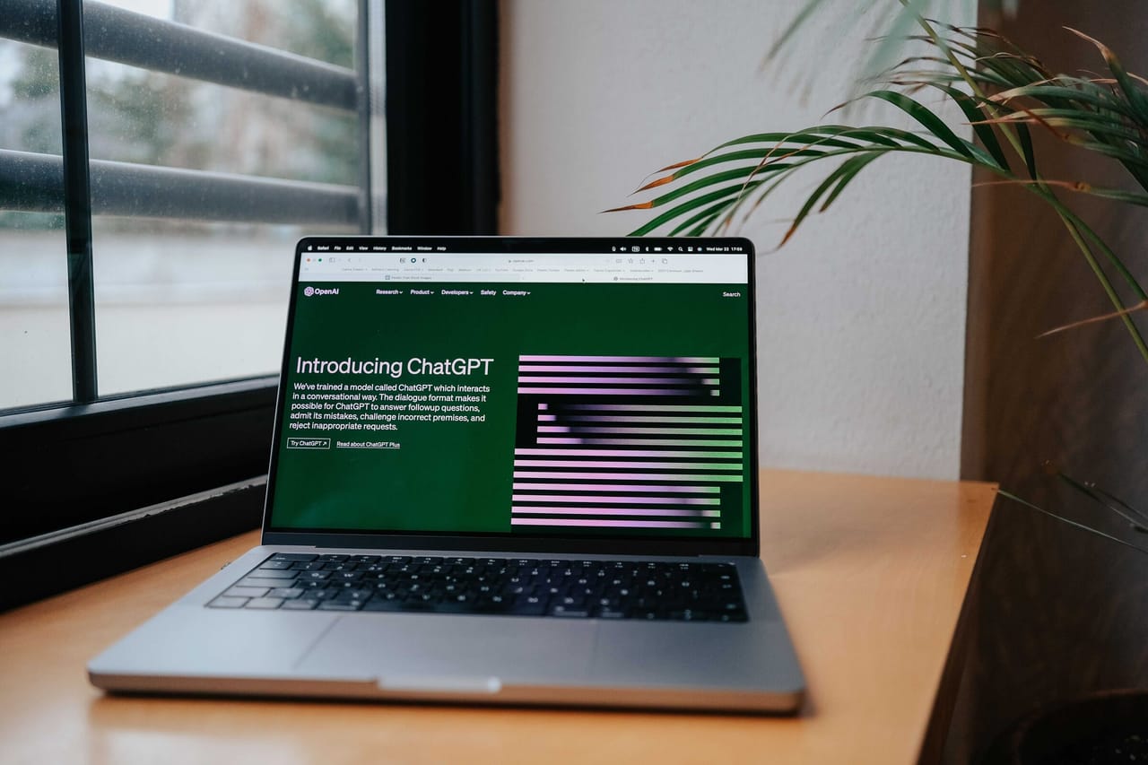 A laptop open on a wooden desk beside a window and a houseplant, showing OpenAI's 'Introducing ChatGPT' announcement page on screen